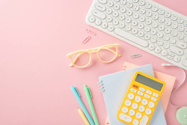 Top view of notepads, pens, glasses, calculator, pc keyboard and other essentials on a pastel pink backdrop.